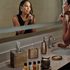 Gold | Woman applying makeup in front of a bathroom mirror with various toiletries on the counter.
