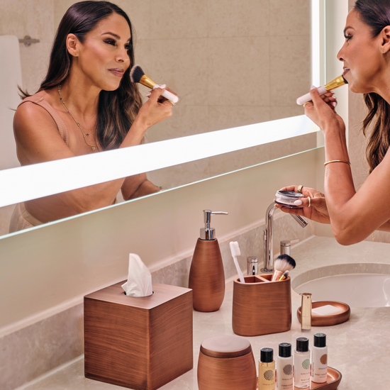 Oil Rubbed Bronze | Woman applying makeup in front of a bathroom mirror with various toiletries on the counter.