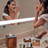 Acacia | Woman applying makeup in front of a bathroom mirror with various toiletries on the counter.