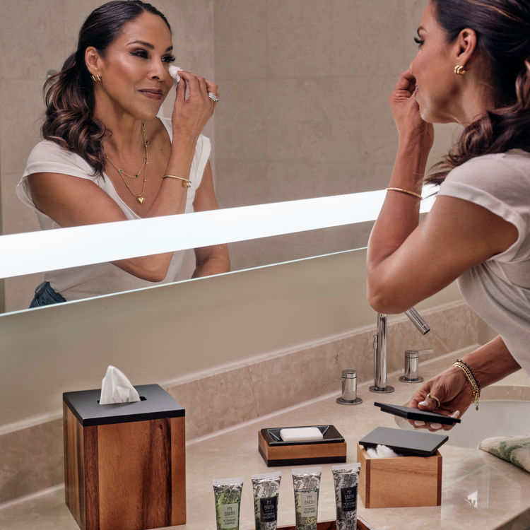 Acacia | Woman applying makeup in front of a bathroom mirror with various toiletries on the counter.