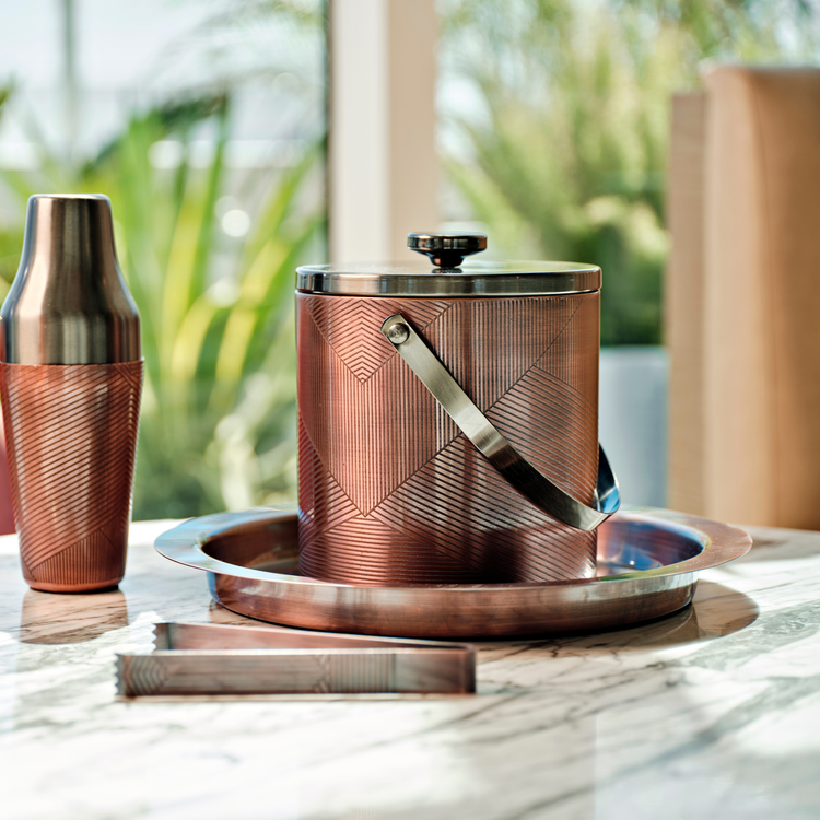 Oil Rubbed Bronze | Copper-themed cocktail shaker and ice bucket set on a marble surface with blurred greenery in the background.